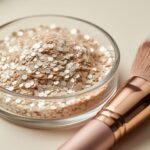Pearly mica shimmer flakes in a clear glass dish with a makeup brush nearby under soft studio lighting.